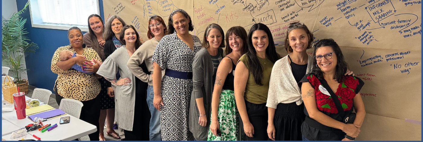 Twelve members of the SAFE Infant Feeding Team standing together, smiling, in front of a wall of butcher paper covered in handwritten planning notes about rapid needs assessments, kits, unity, and informed milk sharing. One team member is holding an infant.